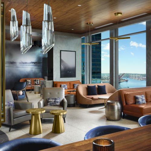 A modern lounge area with luxurious seating, golden accents, large windows showcasing a waterfront view, and elegant lighting fixtures adorn the ceiling.