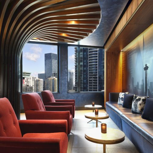 A modern lounge with red armchairs, a curvy wooden ceiling, a cityscape view, and a blue mural wall with cushioned seating and round tables.