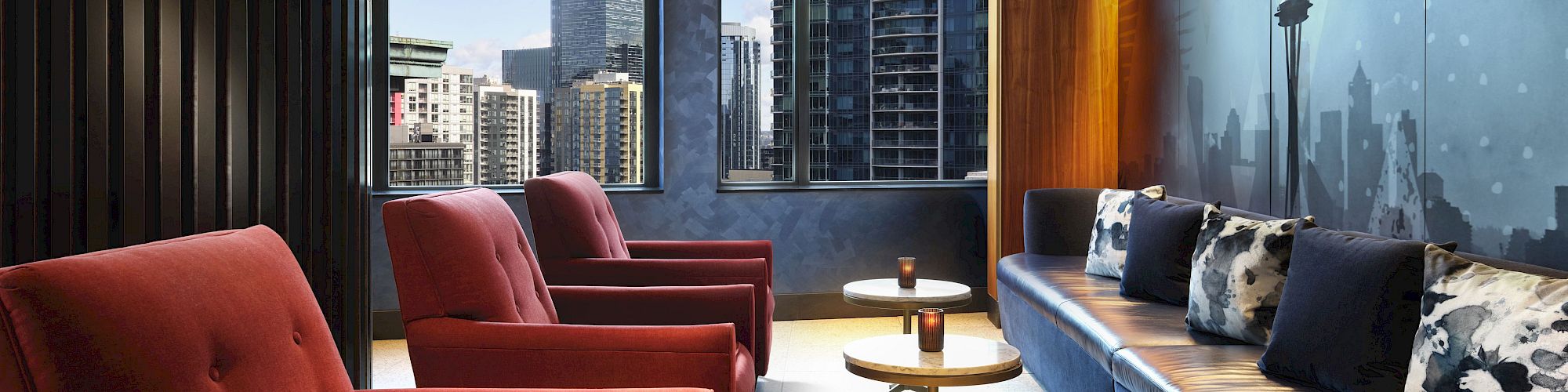 A modern lounge area with red chairs, sleek tables, and a long bench with pillows against a wall featuring a cityscape mural. Ends the sentence.